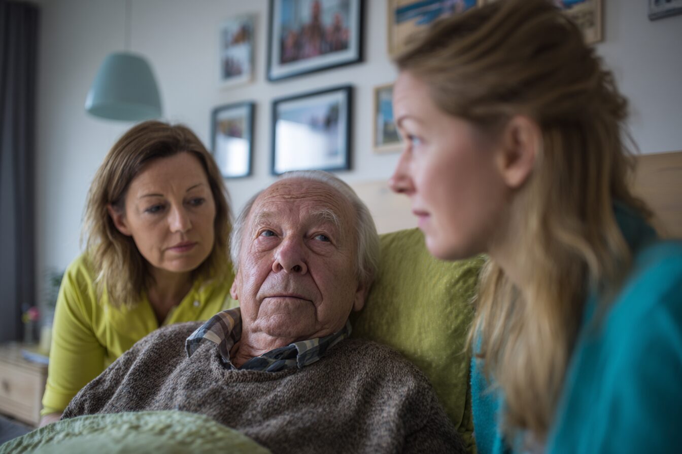 Ein älterer Mann im Bett, umgeben von seiner Tochter und Ehefrau – familiäre Nähe und Palliativpflege zuhause